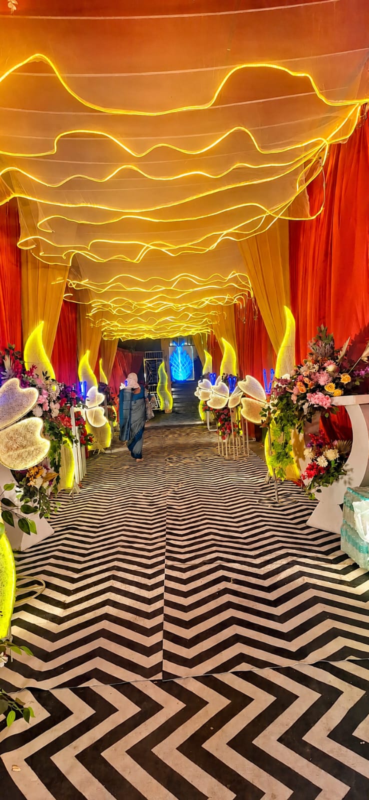 Neon-lit draped ceiling tunnel with chevron floor and floral stands at Maa Durga Palace