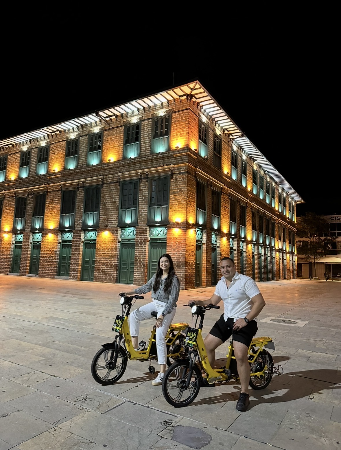 Pareja en bicicletas eléctricas de noche frente a un edificio histórico iluminado de Medellín
