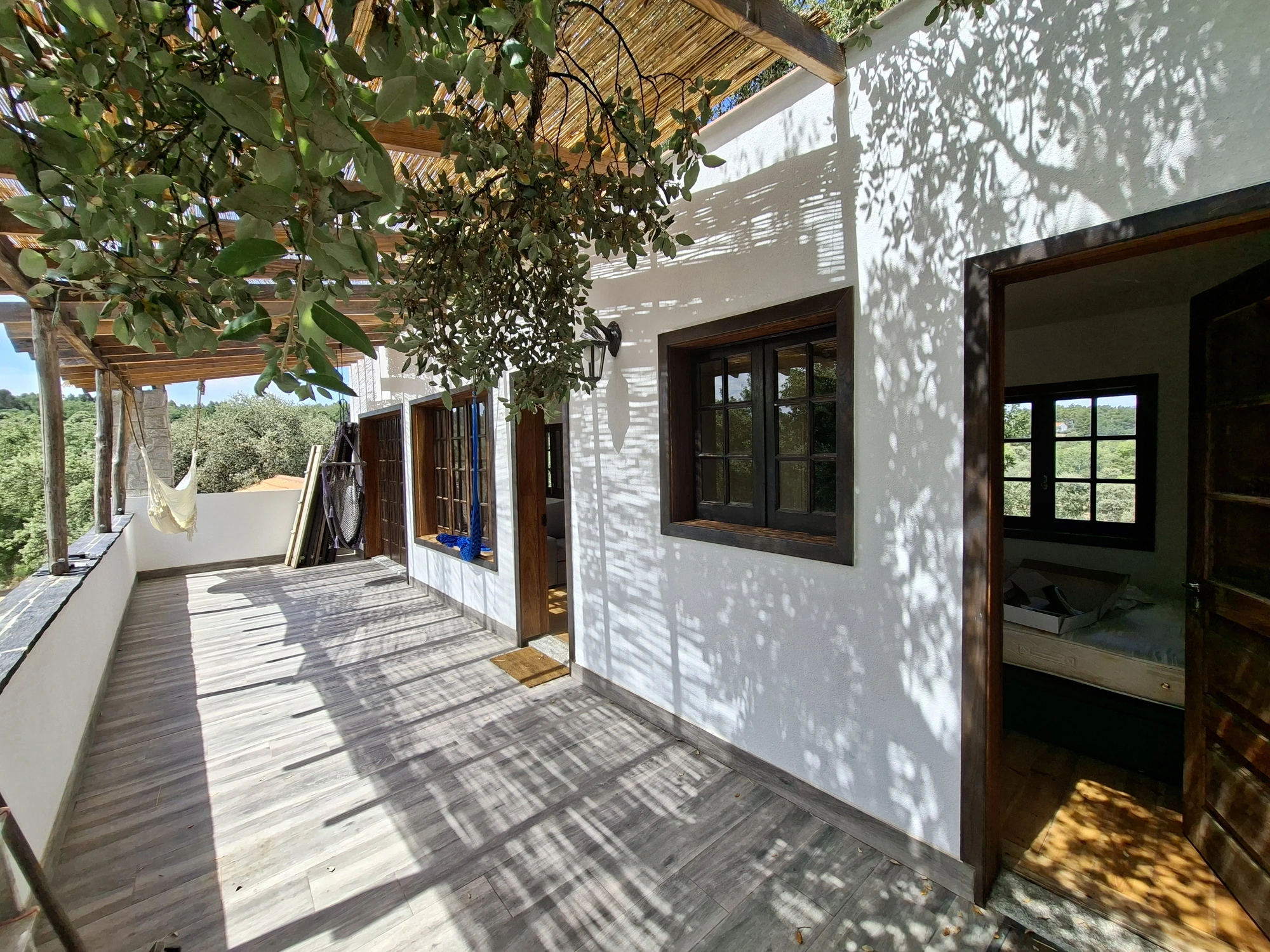 A quiet whitewashed veranda at Limiar under a wooden pergola with a cane roof — soft dappled leaf shadows on the walls, open doorways revealing bedrooms inside, a suspended hammock chair on the left and the deck stretching into the treeline
