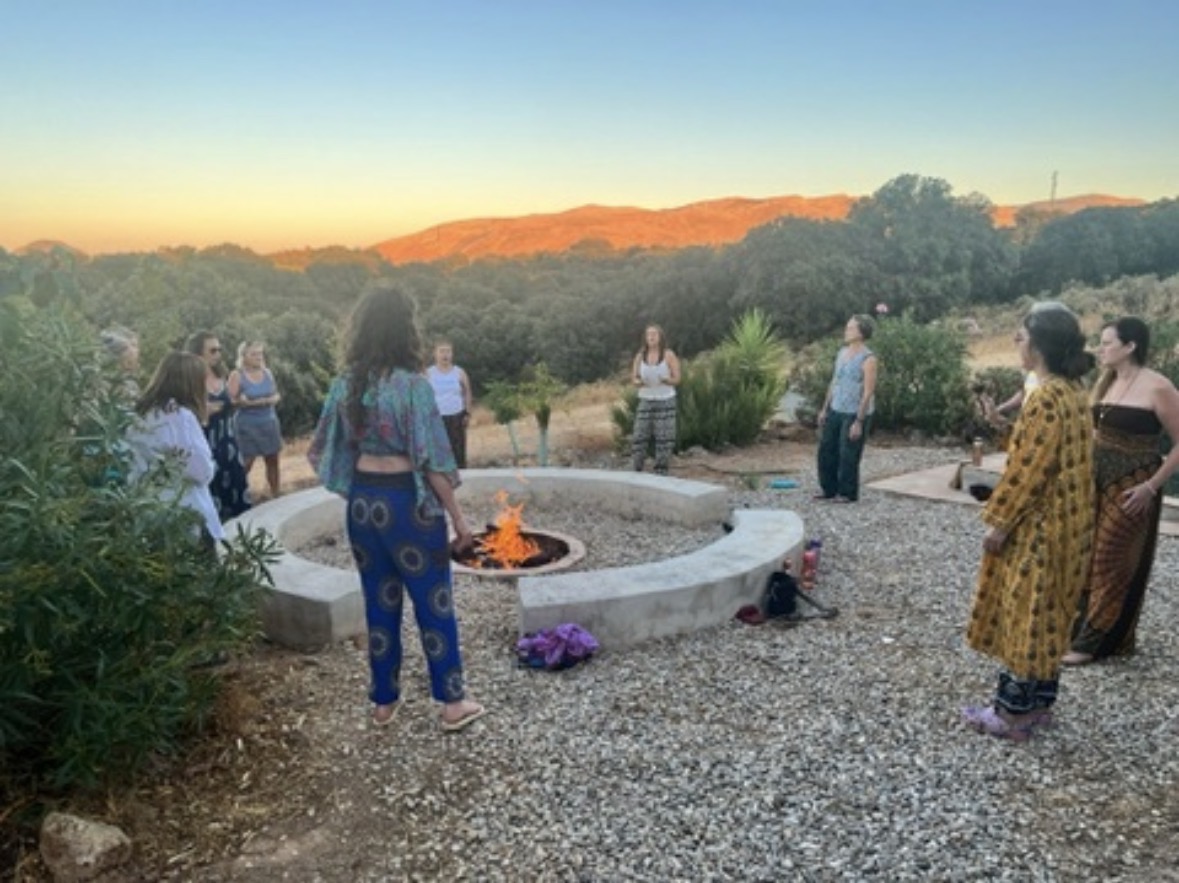 Women's circle gathered around an outdoor stone fire pit at sunset, with mountains glowing orange in the distance and women standing in a circle holding space together