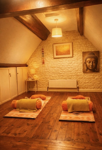 The warm loft yoga room inside Annelie's Grade II listed cottage in Frome with stone wall, Buddha artwork, chakra hanging, sunset painting and two yoga mats with bolsters under soft lamp light