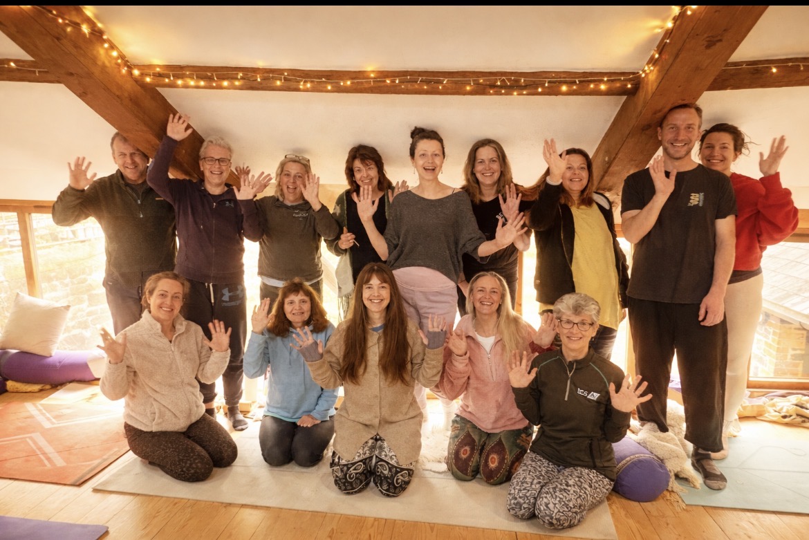 Joyful retreat group of around 16 participants smiling and waving in the warm loft room with fairy lights and wooden beams during an Annelie Lily retreat