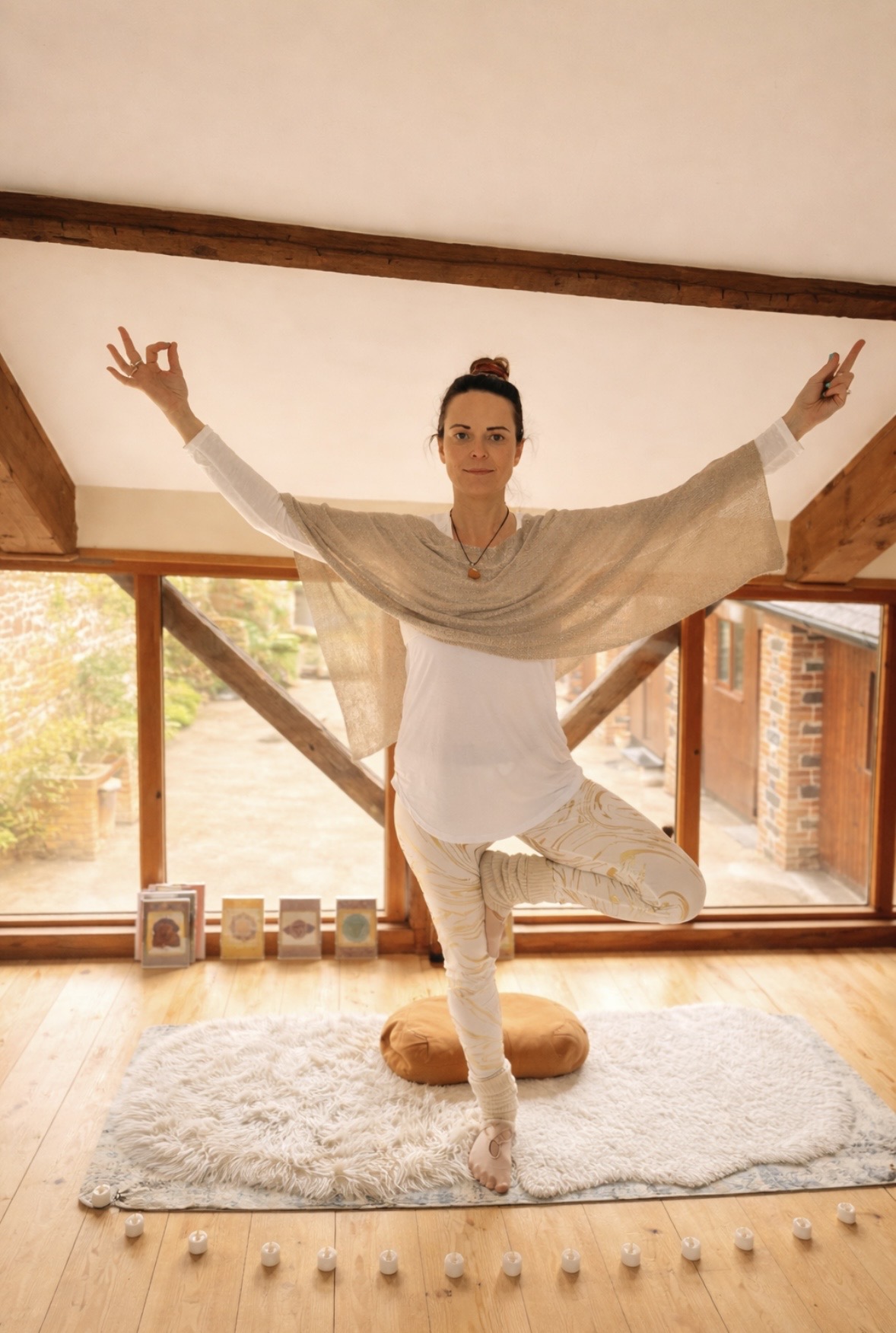 Annelie Lily standing in tree pose with arms raised in mudra inside the warm loft yoga room of her Grade II listed cottage in Frome, with wooden beams, candles and golden window light