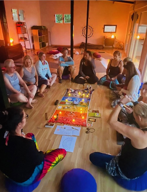 Women seated together in a circle around a colourful chakra mandala spread with candles, oracle cards and reflection journals in a warm sunlit studio