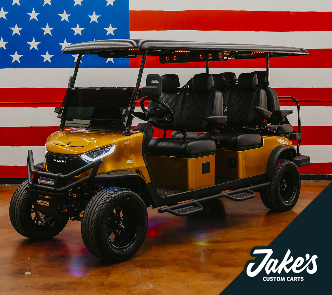 A custom gold-painted KANDI 6-Pro 6-passenger lifted golf cart with extensive custom work and accessories, photographed in the Jake's Custom Carts Tampa showroom in front of the painted American flag wall