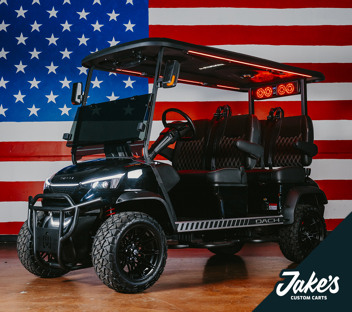 A blacked-out DACH 6-passenger lifted golf cart with red LED accent lighting and quad-speaker overhead audio, photographed in the Jake's Custom Carts Tampa showroom in front of the shop's painted American flag wall