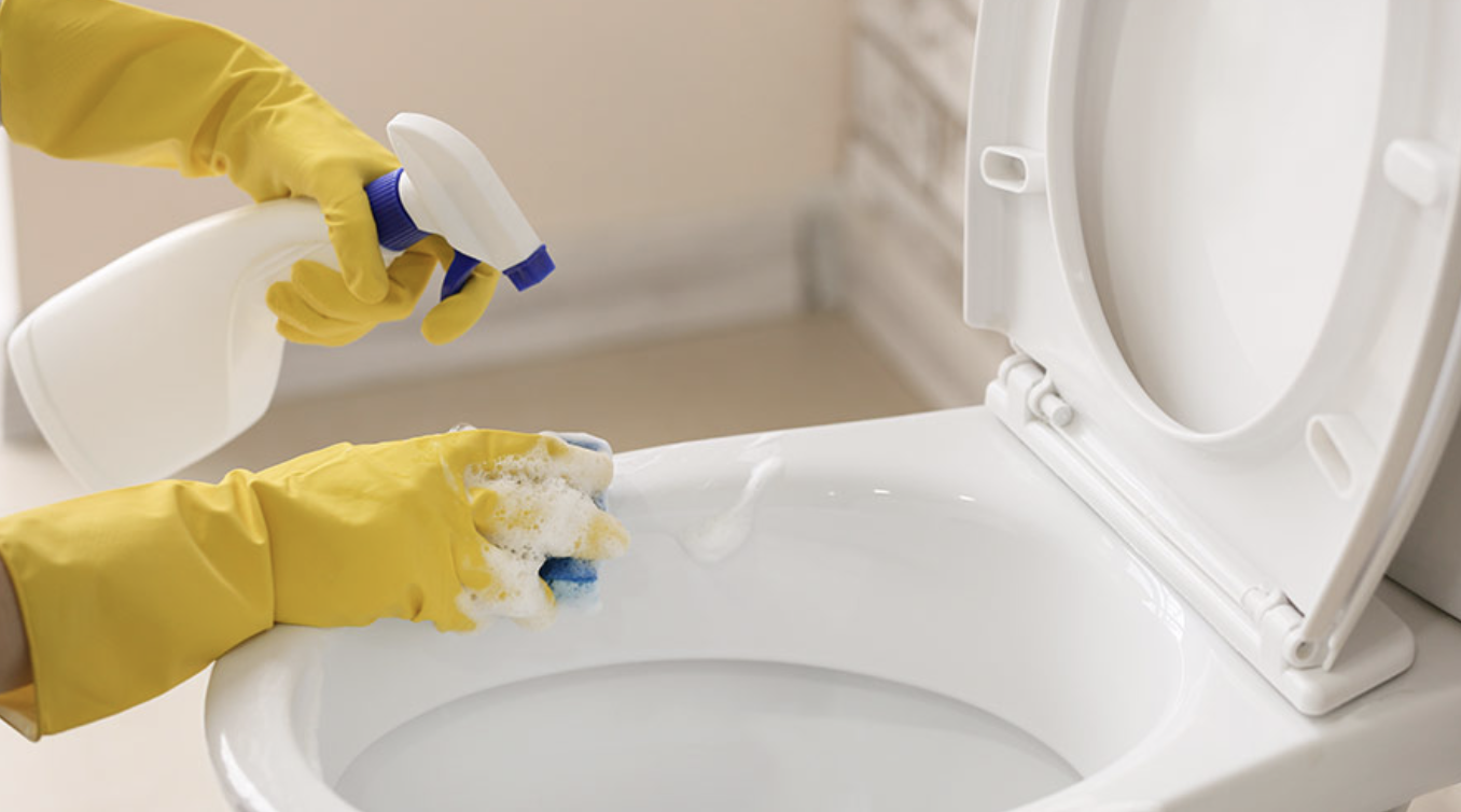 Cleaning technician in yellow rubber gloves spraying disinfectant and scrubbing a commercial toilet bowl