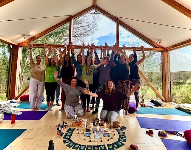 Yoga retreat group celebrating with arms raised in glass pavilion with sacred mandala altar at The Station, Frome