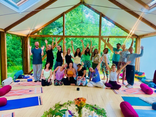 Joyful yoga retreat group with arms raised in The Station glass pavilion surrounded by green trees in Frome