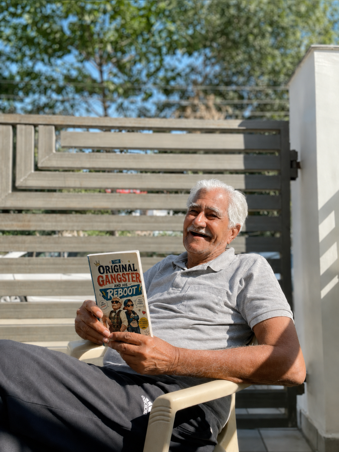 A grandfather sitting on a sunlit balcony, holding a custom Keepsake Storybook titled 'The Original Gangster and His Reboot'