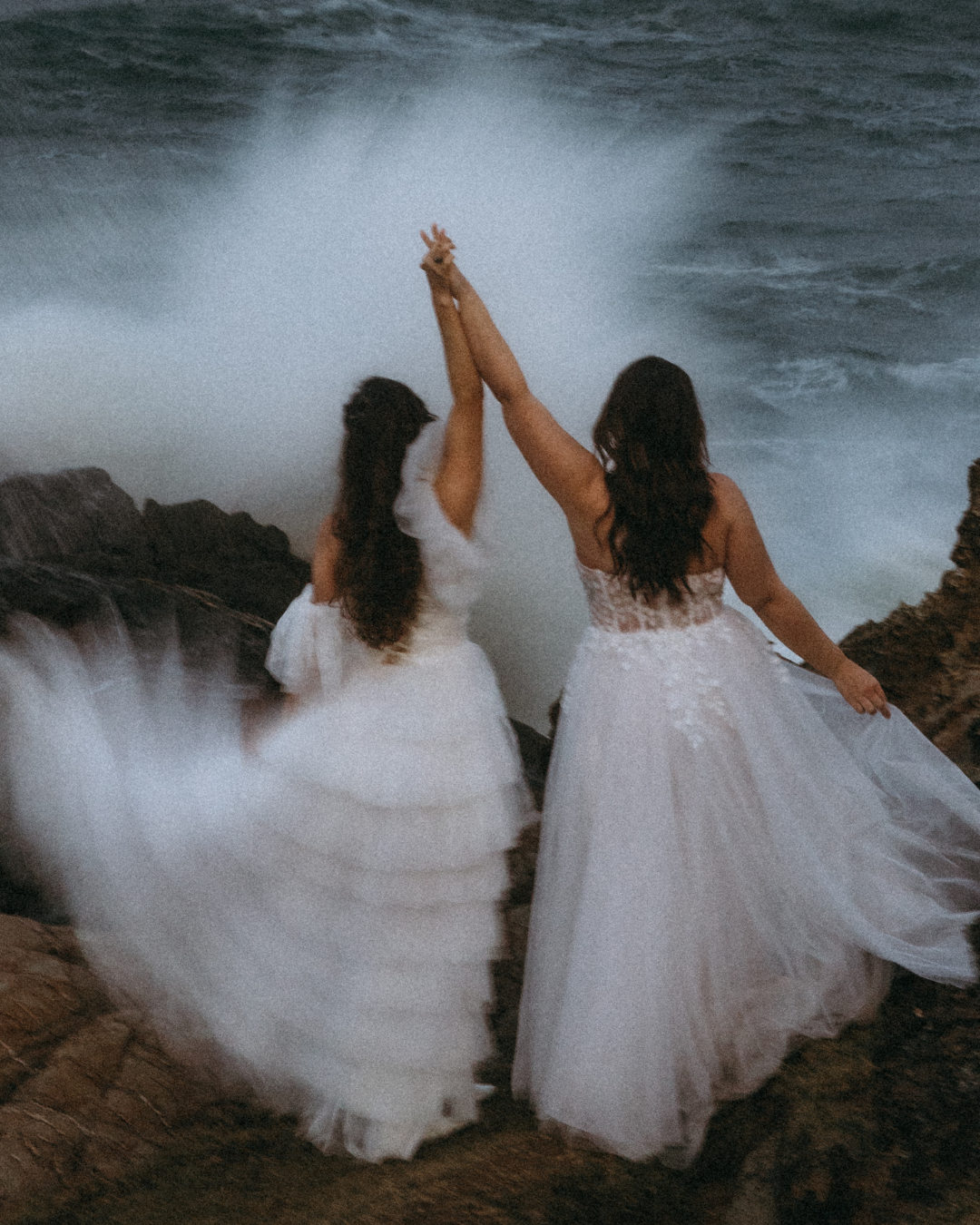 Two brides celebrating on a coastal cliff, hands raised together
