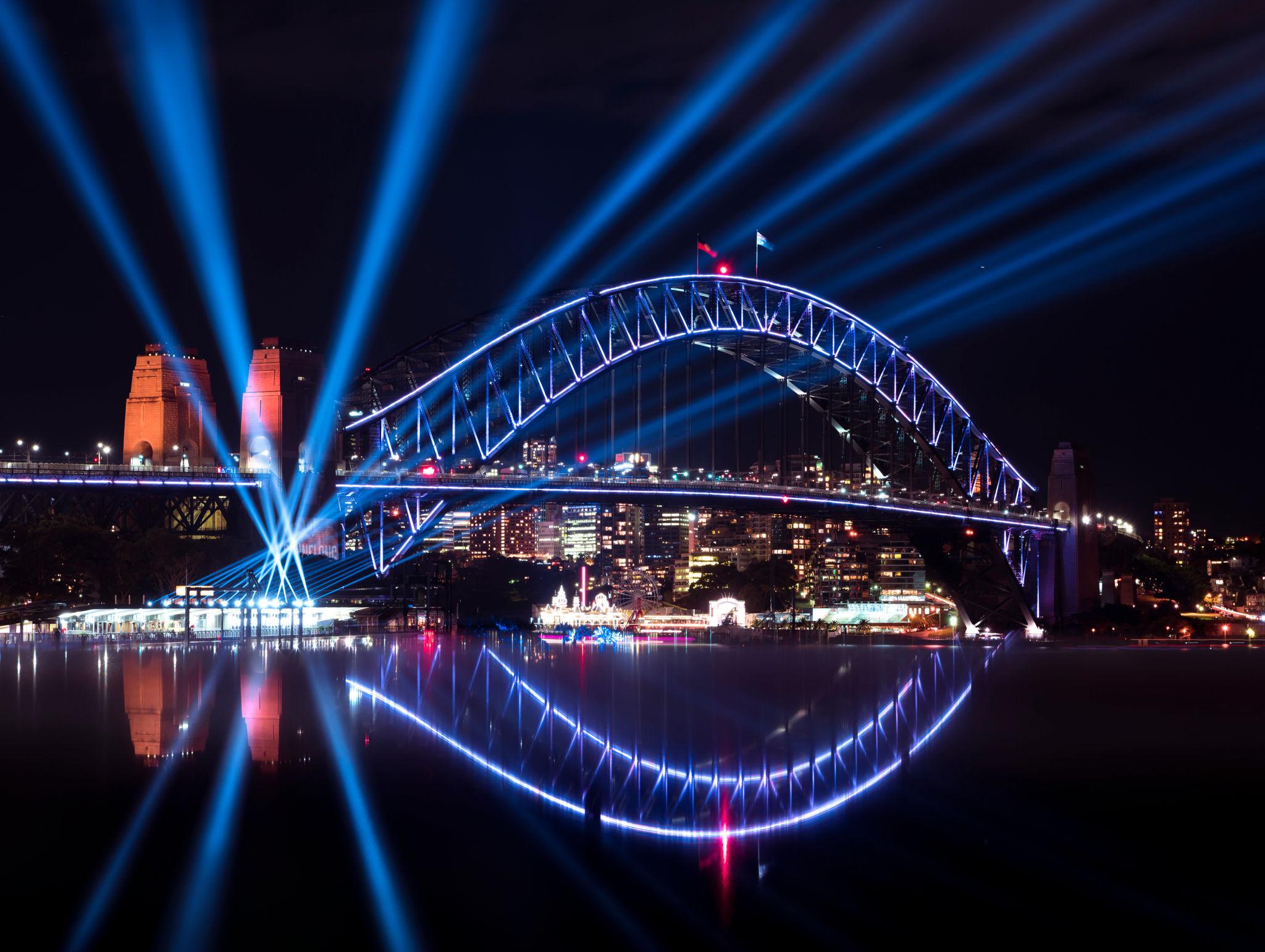 Vivid Sydney cruise · Harbour Bridge lit in blue laser beams reflecting on the water · view from the Magic