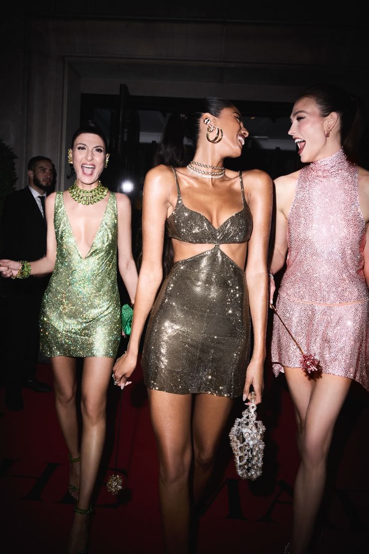 Three women laughing together in shimmering luxury evening dresses at a glamorous event