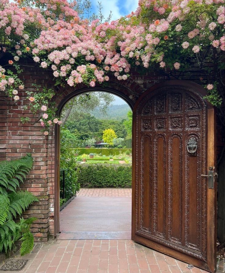 An arched garden gate with climbing pink roses, the carved wooden door slightly ajar, opening into a sunlit garden beyond
