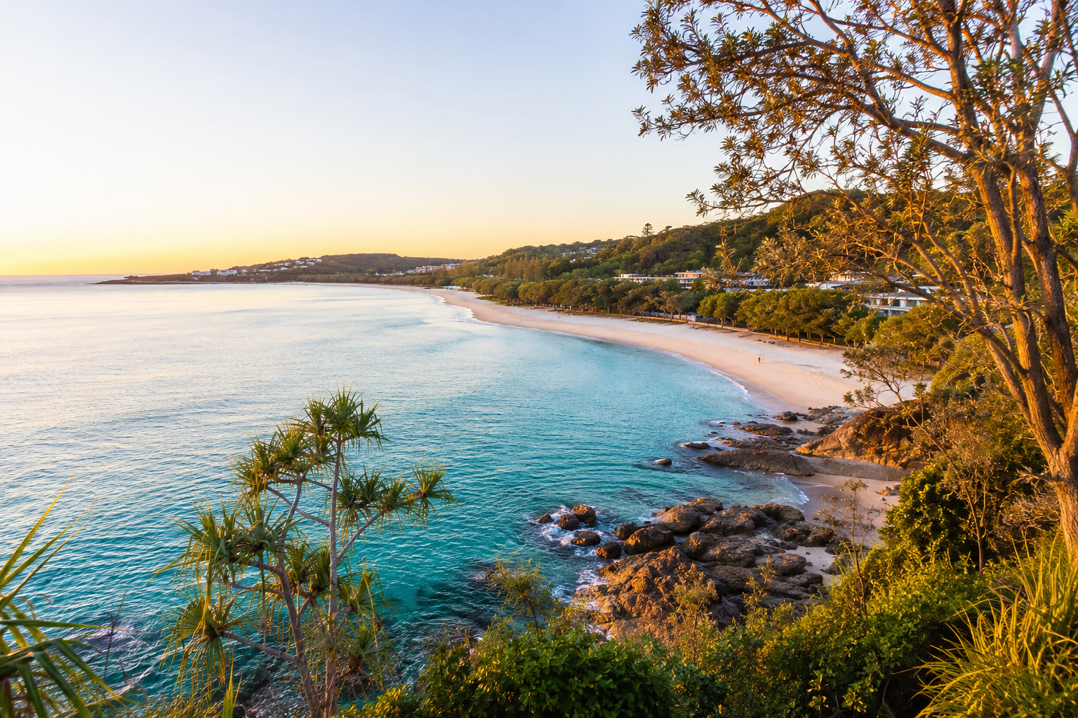 Golden-hour view of Noosa Heads main beach from the headland, with calm turquoise water, white sand, coastal vegetation in the foreground and the Noosa National Park escarpment beyond