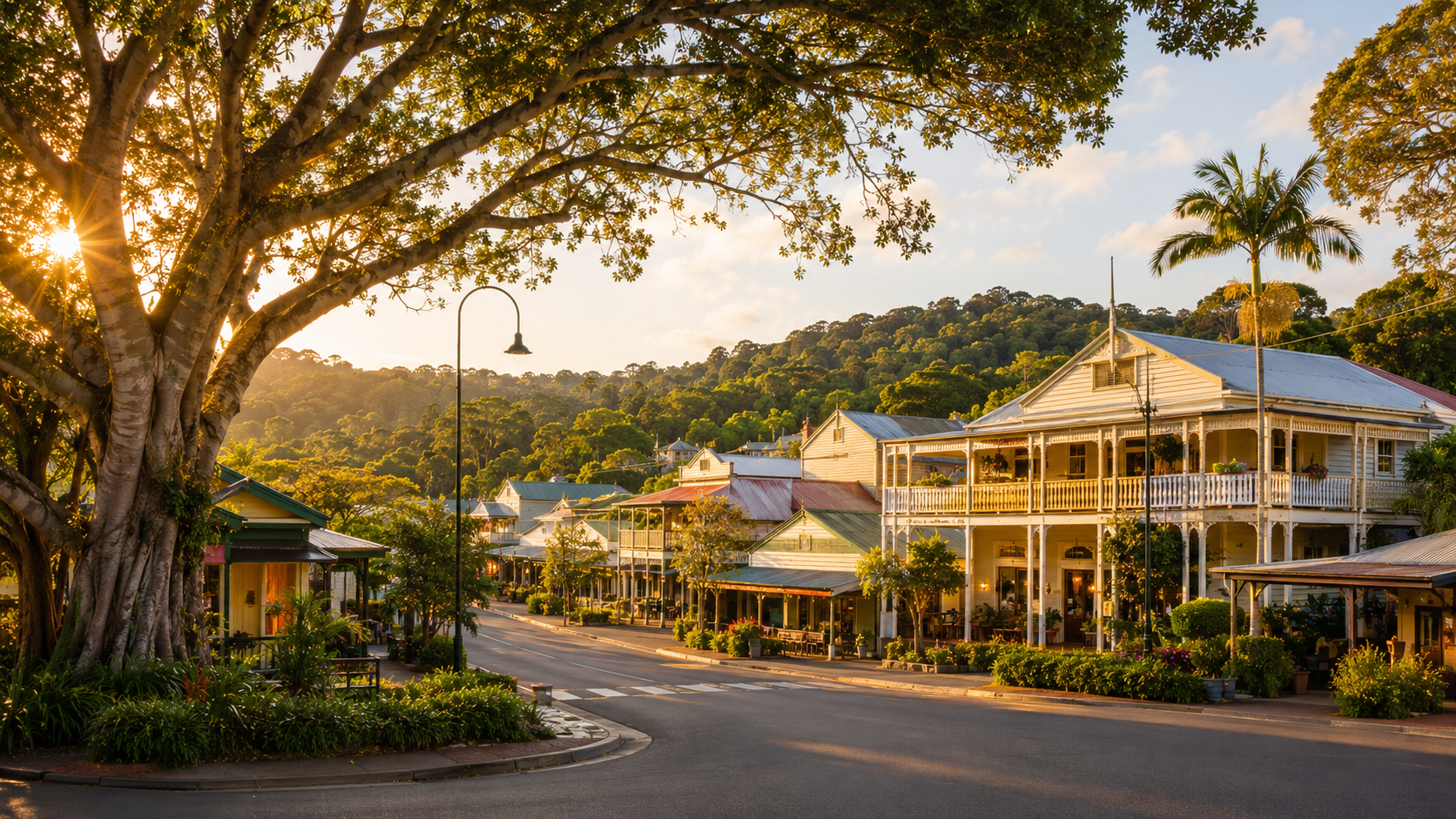 Golden-hour view of Eumundi's heritage main street with verandahed Queenslander shopfronts, a large shade tree, palms and the hinterland ridge rising behind the township