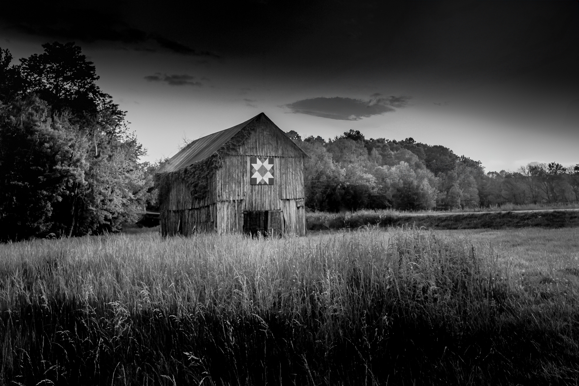 Black-and-white photograph of a quilt barn beside a country road and open field