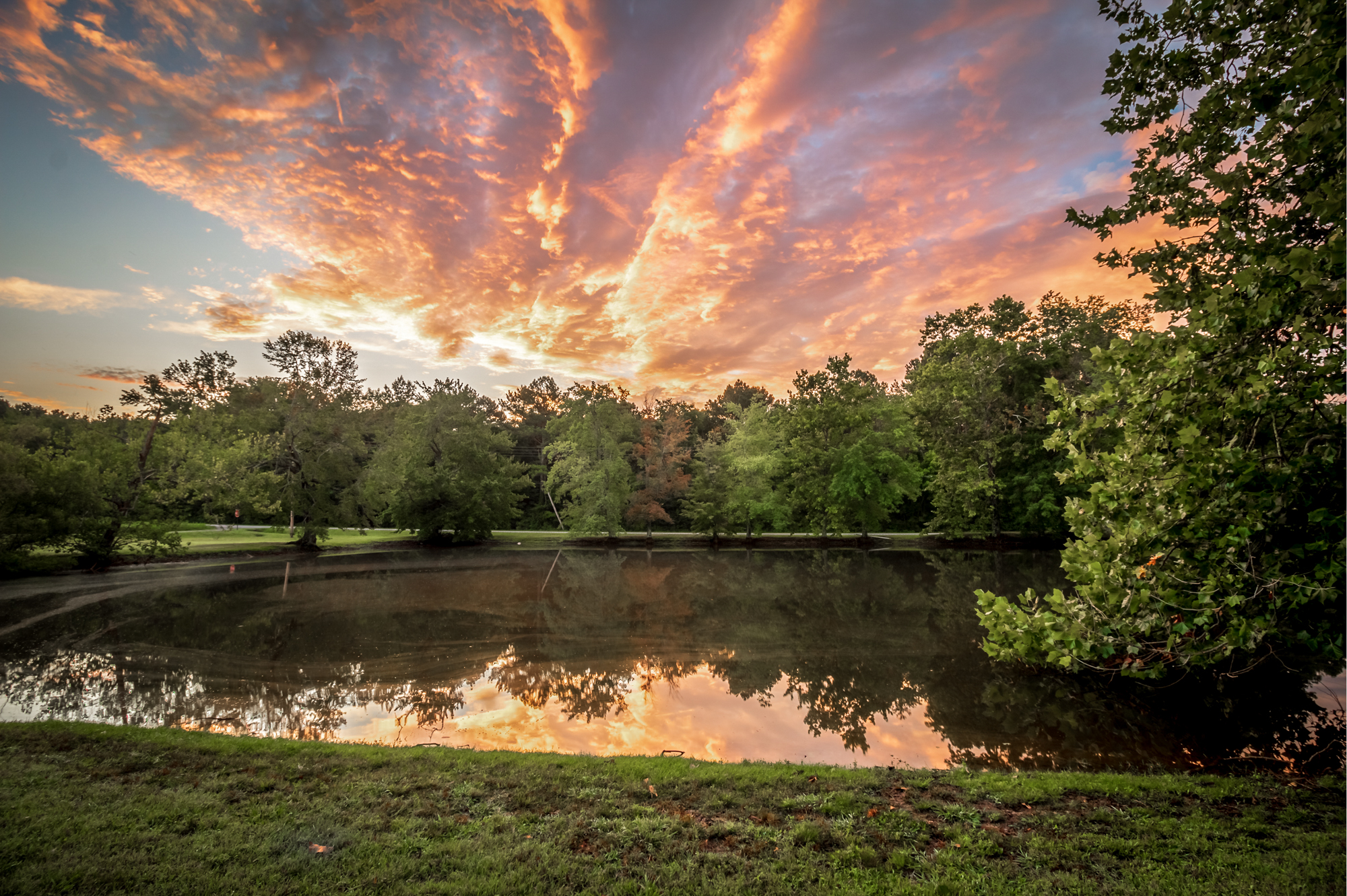 Color photograph of a dramatic glowing sky above trees and a lake reflection
